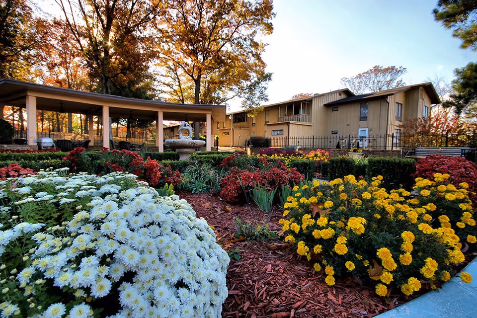 A vibrant garden with blooming white, yellow, and red flowers in front of a beige two-story building with balconies. There is a covered outdoor seating area with a fountain in the center, surrounded by trees with autumn foliage.