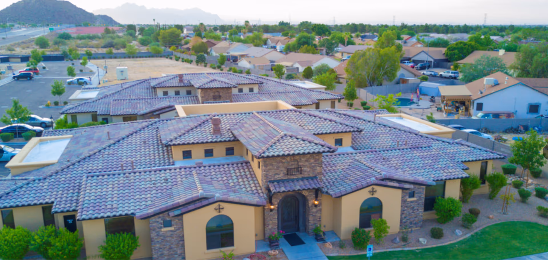 Aerial view of a large residential building with a tiled roof and stone accents, surrounded by greenery and neighboring houses. The building has a prominent entrance with an arched doorway and is part of a suburban neighborhood with visible streets, parked cars, and distant mountains.