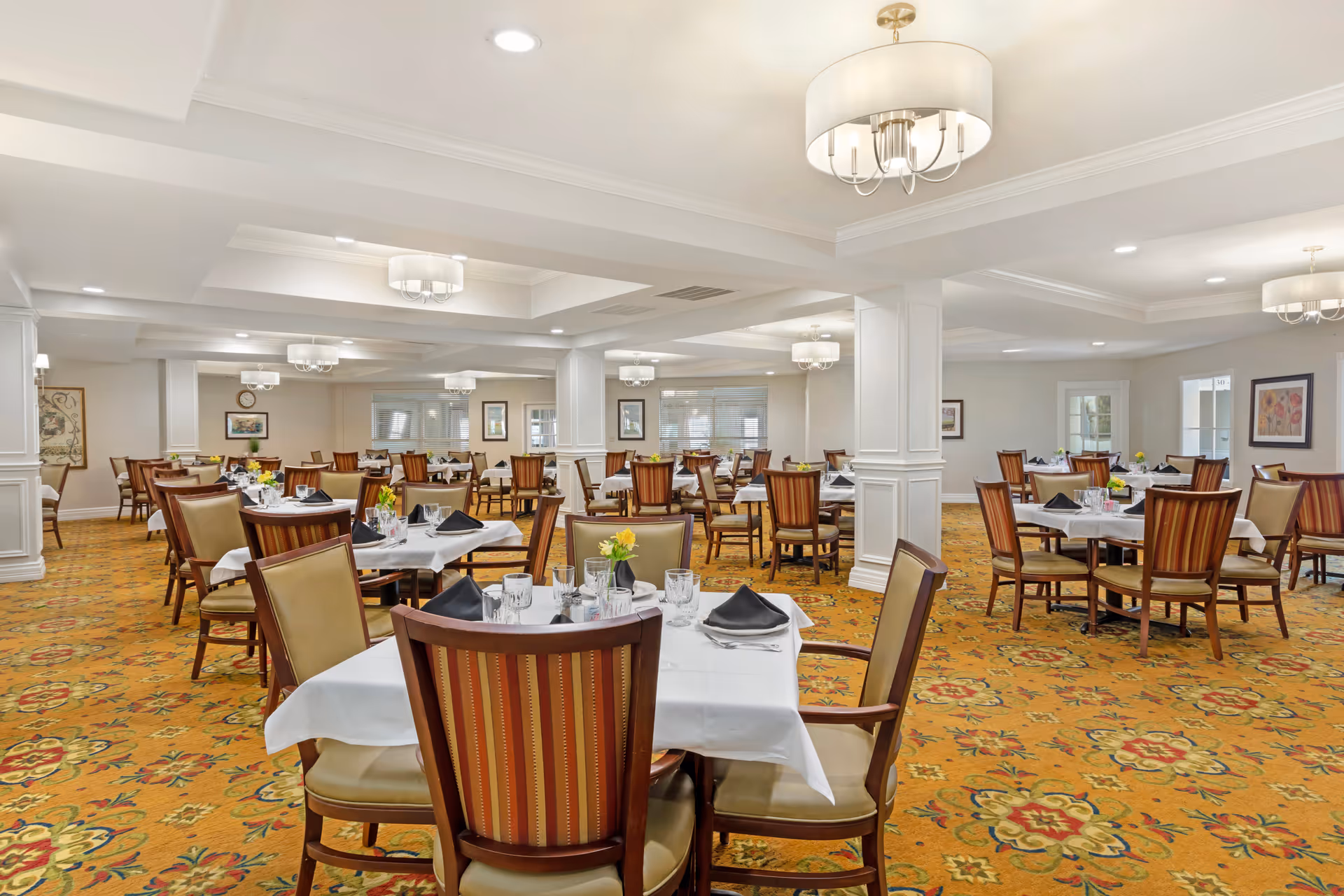 A spacious dining room with multiple tables covered in white tablecloths, each set with glassware, silverware, black napkins, and small yellow flower centerpieces. The room features patterned carpet, white walls, several columns, and modern ceiling light fixtures.