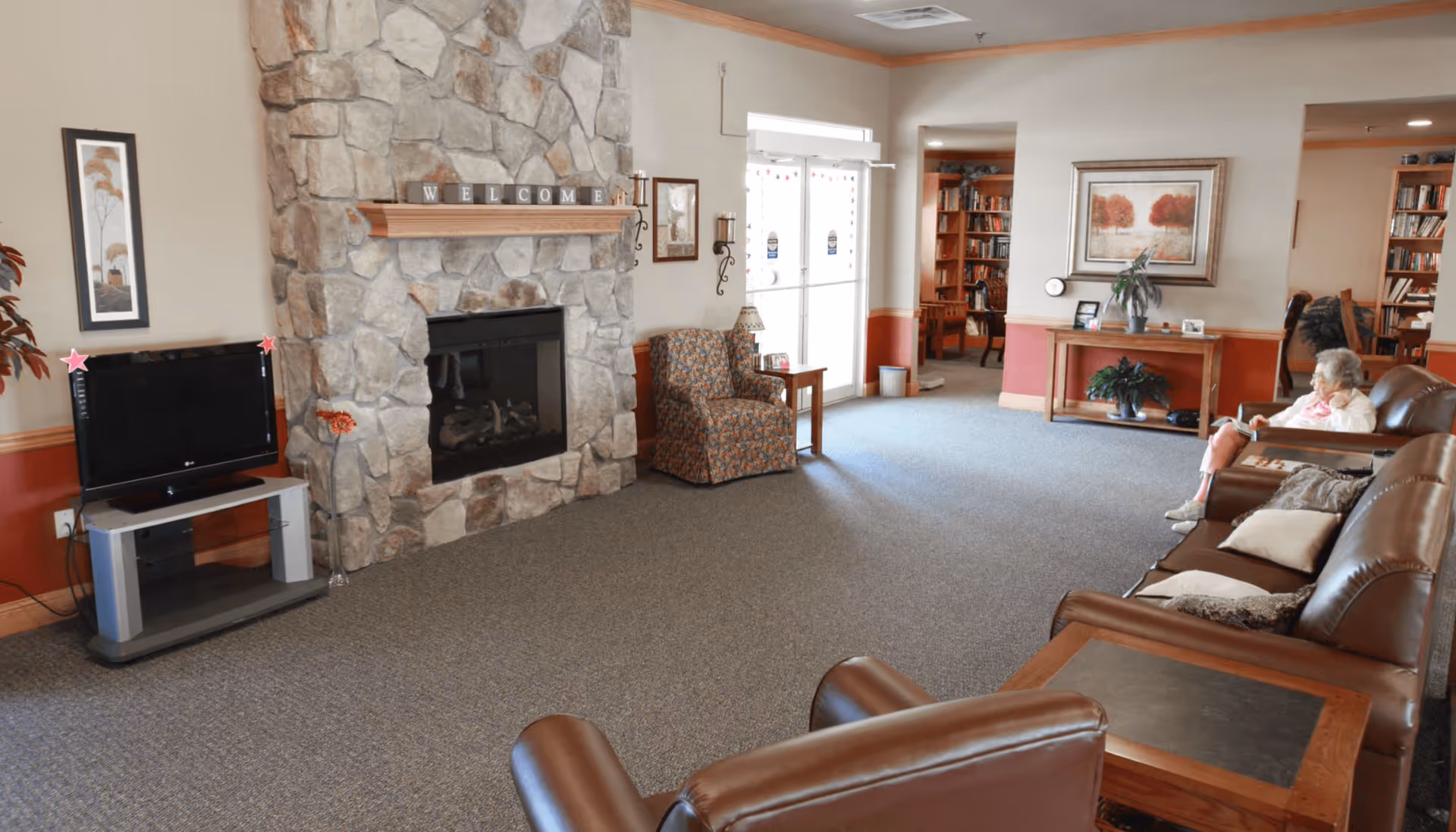 A cozy assisted living facility common area with a stone fireplace, a flat-screen TV on a stand, a floral armchair, and leather sofas. An elderly woman is sitting on one of the sofas reading a book. The room has carpeted floors, framed artwork on the walls, and a table with plants and picture frames near the entrance.