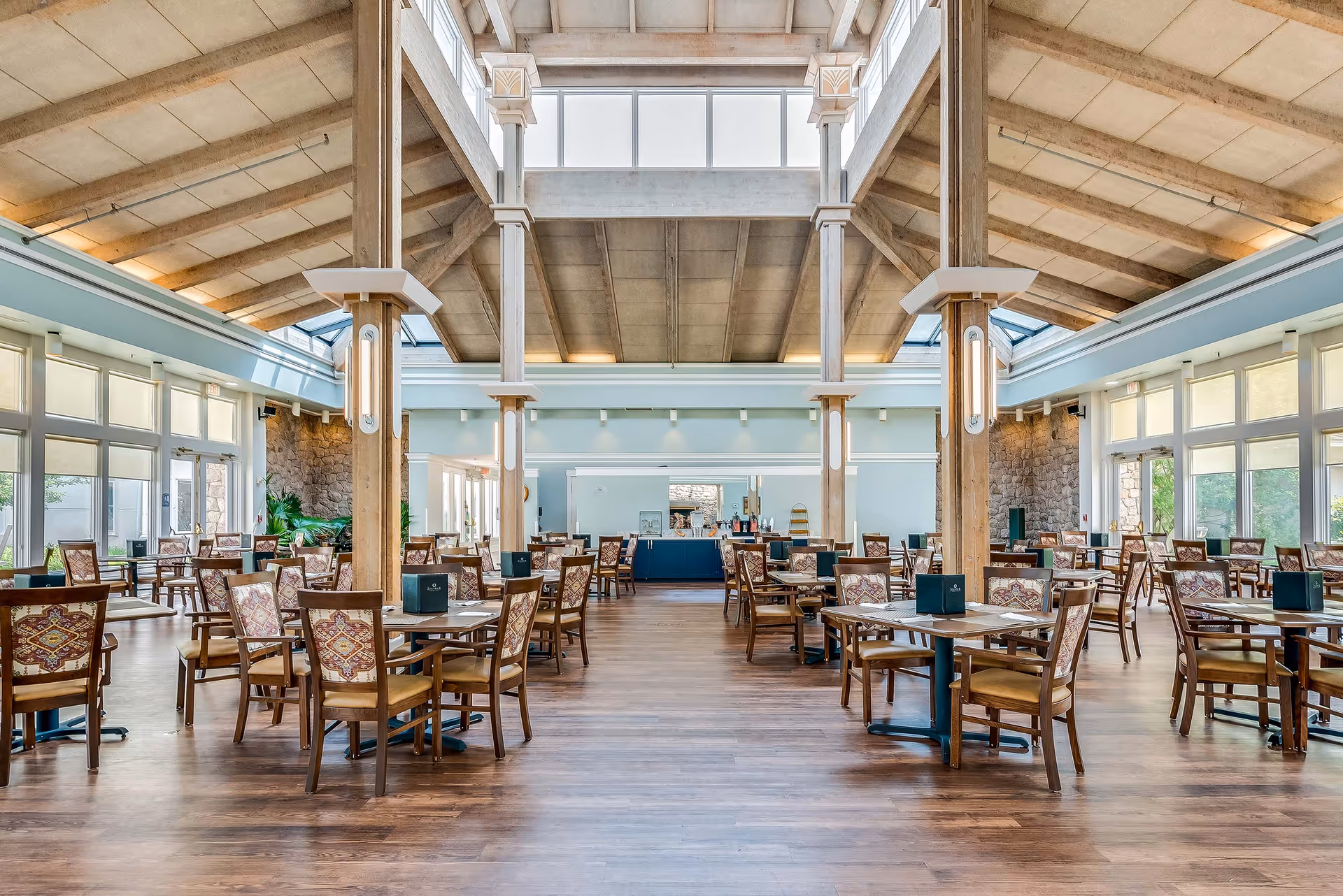 Spacious dining room with many wooden tables and patterned chairs under a high beamed ceiling and skylights.