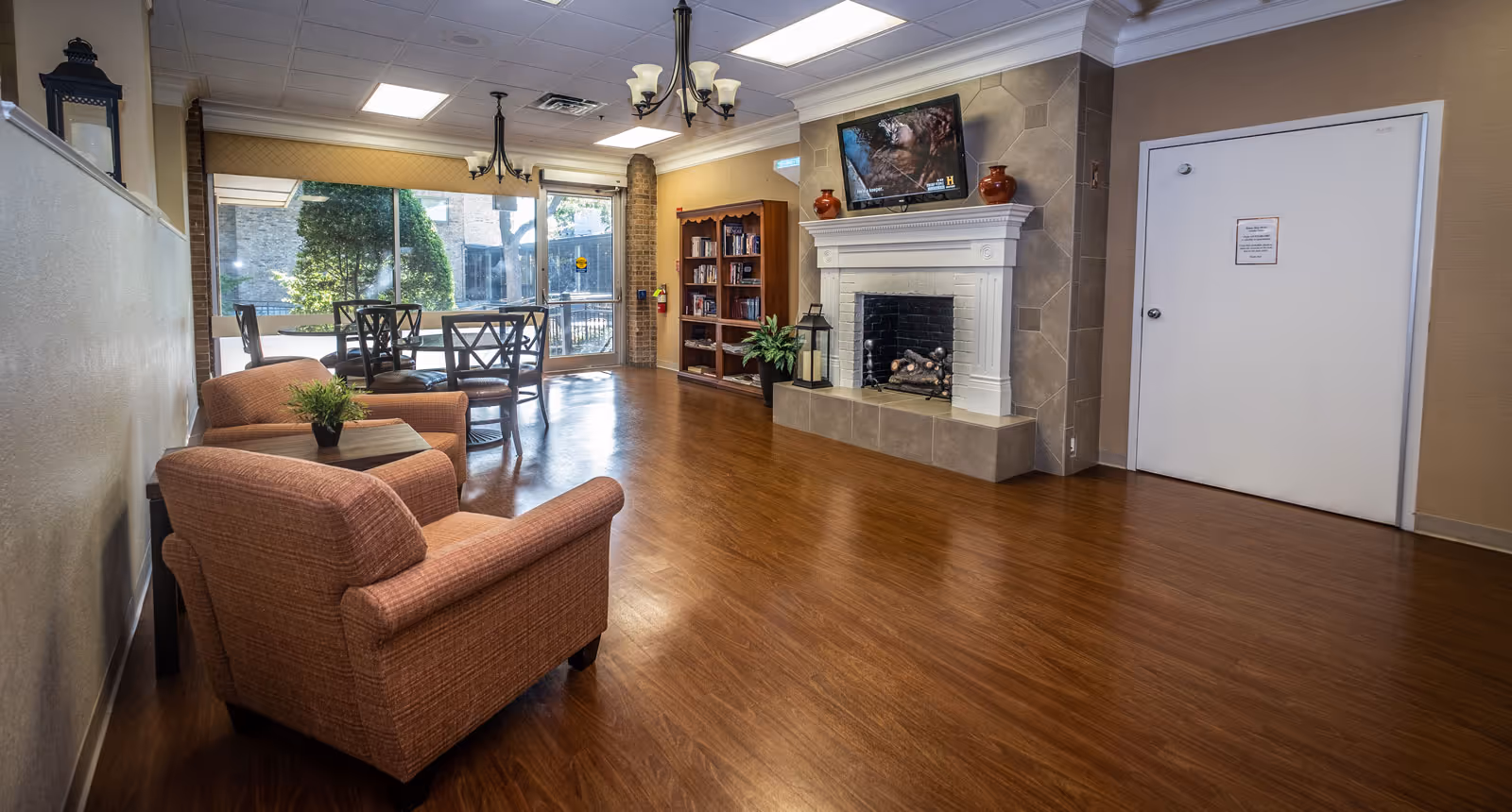 A cozy common area in a senior living facility featuring two upholstered armchairs with a small table and plant between them, a round table with four chairs near large windows letting in natural light, a bookshelf filled with books, a fireplace with decorative vases on the mantel, and a wall-mounted television above the fireplace.