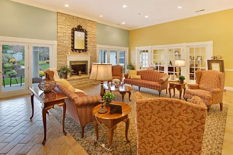 A cozy senior living facility common area with patterned upholstered armchairs and sofas arranged around a wooden coffee table and side tables. The room features a stone fireplace with a decorative mirror above it, large windows and glass doors leading to a garden outside, and soft lighting from table lamps. The walls are painted in soft green and yellow tones, and the floor is covered with a patterned rug and wood-look tiles.