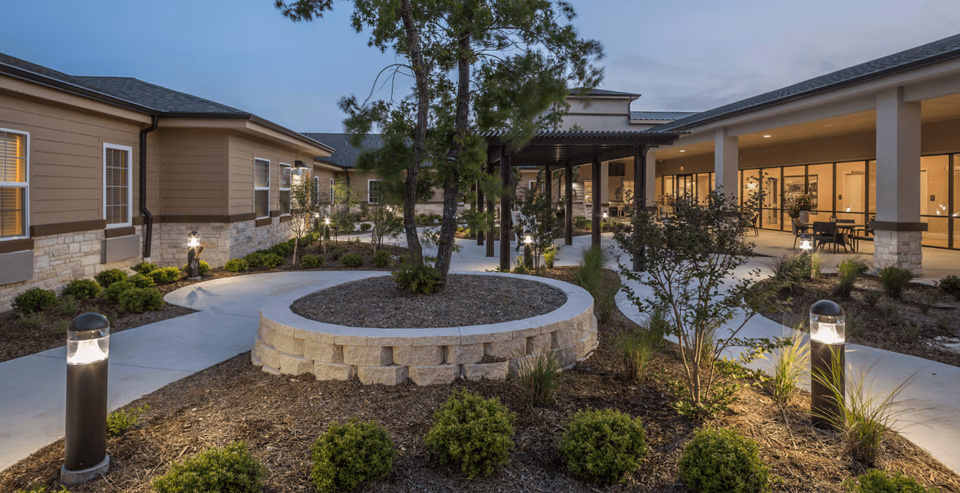 Outdoor courtyard area at dusk with landscaped garden beds, small trees, shrubs, and modern pathway lights. Surrounding buildings with large windows and covered walkways are visible.