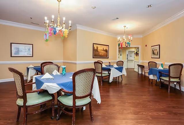Dining room with several tables covered with blue and white tablecloths, each set with napkins and small decorative vases. The room has wooden flooring, beige walls with white trim, framed artwork, and chandeliers decorated with colorful hanging ornaments.