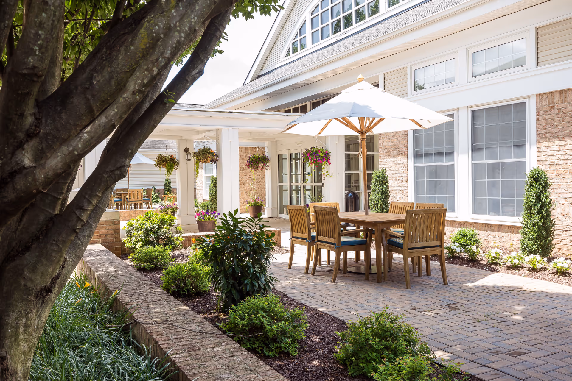 Outdoor patio area at Copper Ridge featuring a wooden table with six chairs and a large white umbrella. The patio is paved with bricks and surrounded by well-maintained landscaping including bushes, flowers, and hanging flower pots. The building exterior is visible with large windows and light-colored siding.