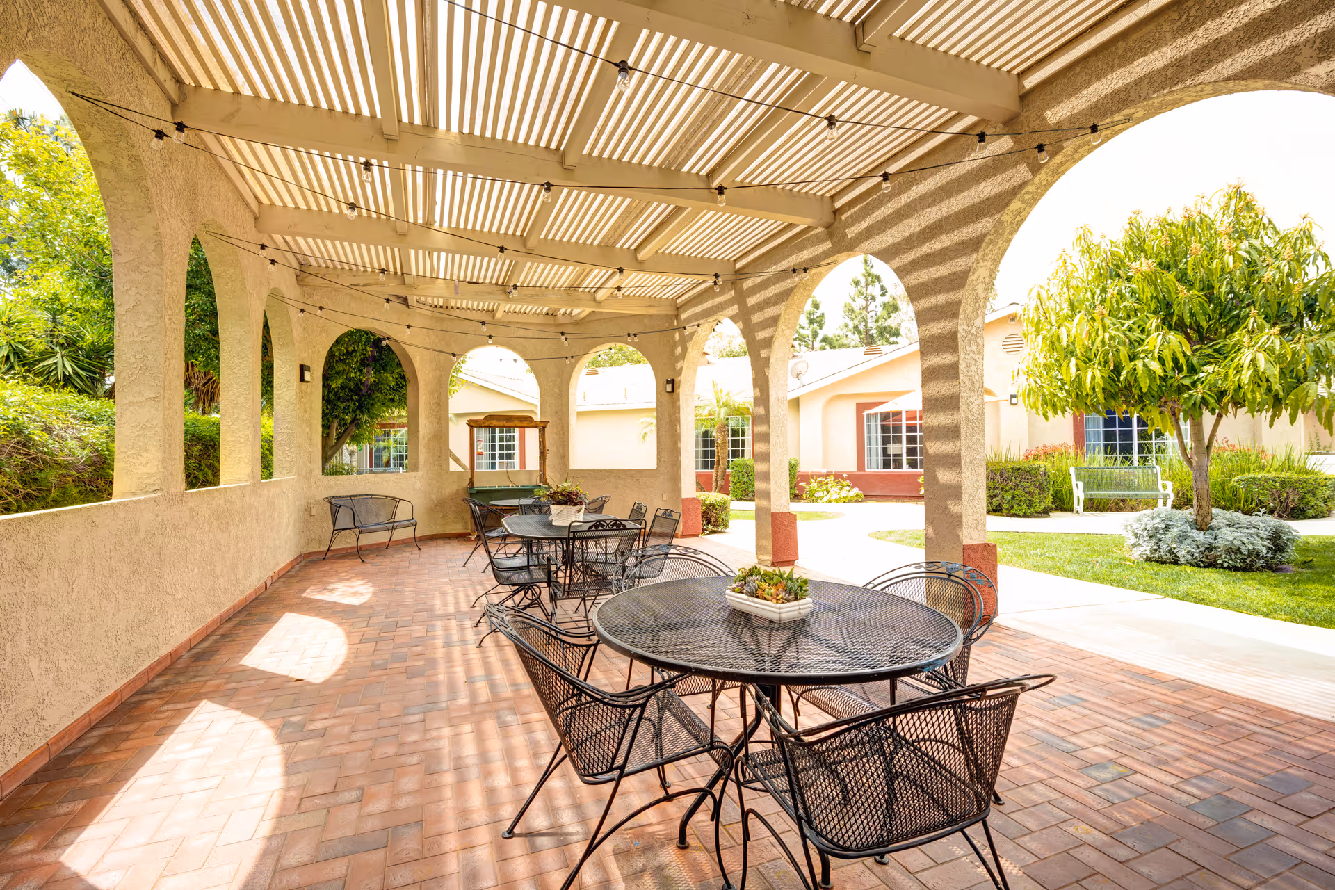 Outdoor covered patio area with a slatted roof casting striped shadows on the brick floor. Several black metal tables and chairs are arranged under the patio. The area is surrounded by beige stucco walls with arched openings, and beyond the patio, there is a well-maintained garden with green grass, bushes, and trees. Residential buildings with windows are visible in the background.