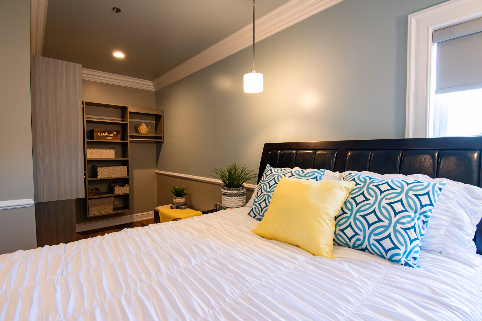 Well-lit bedroom with a large bed topped with blue patterned and yellow pillows, built-in shelving, potted plants, and a hanging pendant light.