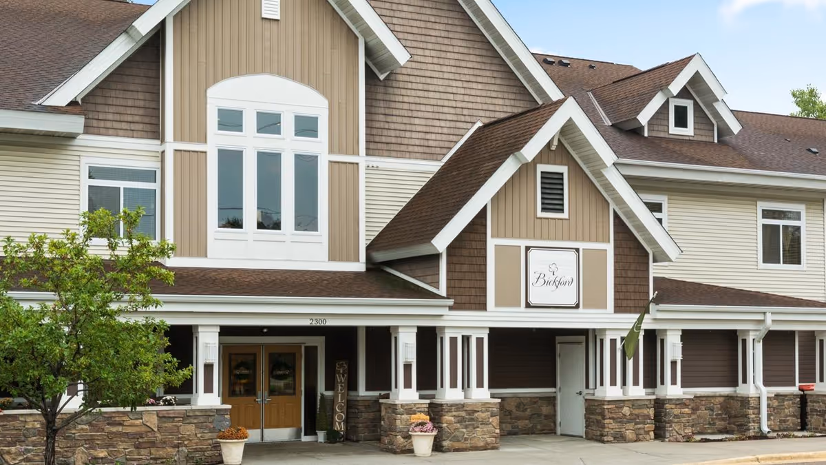 Exterior front view of Bickford of Maplewood senior living facility showing a two-story building with a combination of stone and wood siding, a welcoming entrance with double doors, and a sign displaying the facility name.