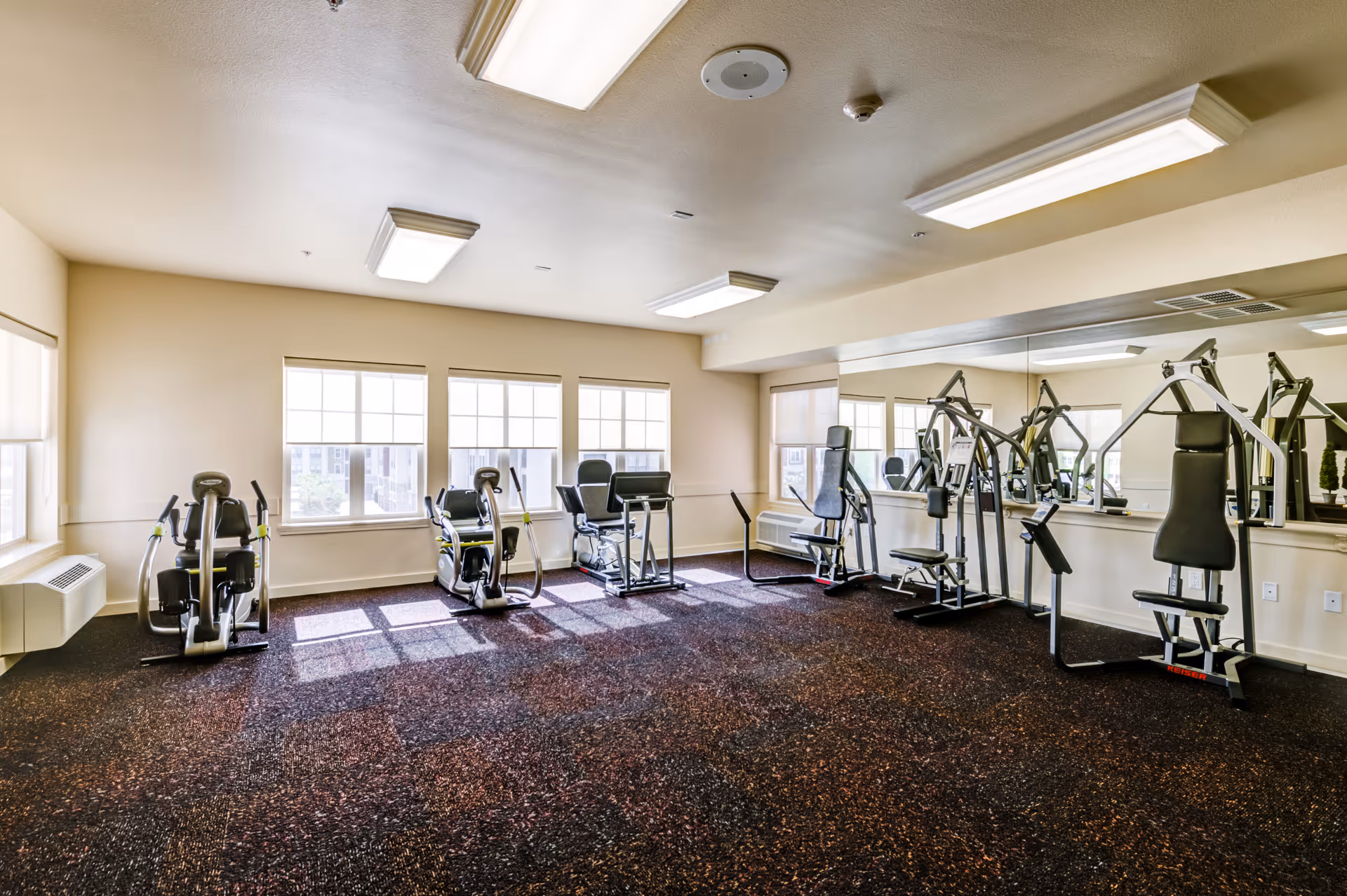 A bright fitness room with exercise machines including stationary bikes and weight machines, large windows letting in natural light, and a wall covered with mirrors.