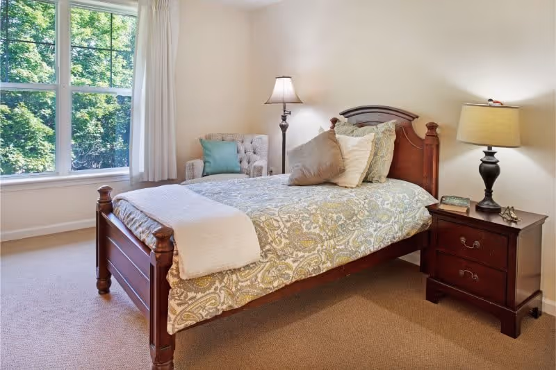 A cozy bedroom with a wooden bed frame, patterned bedspread, and multiple pillows. There is a wooden nightstand with a lamp and a book on it. A cushioned armchair with a green pillow is placed near a large window with white curtains, letting in natural light and showing green trees outside.