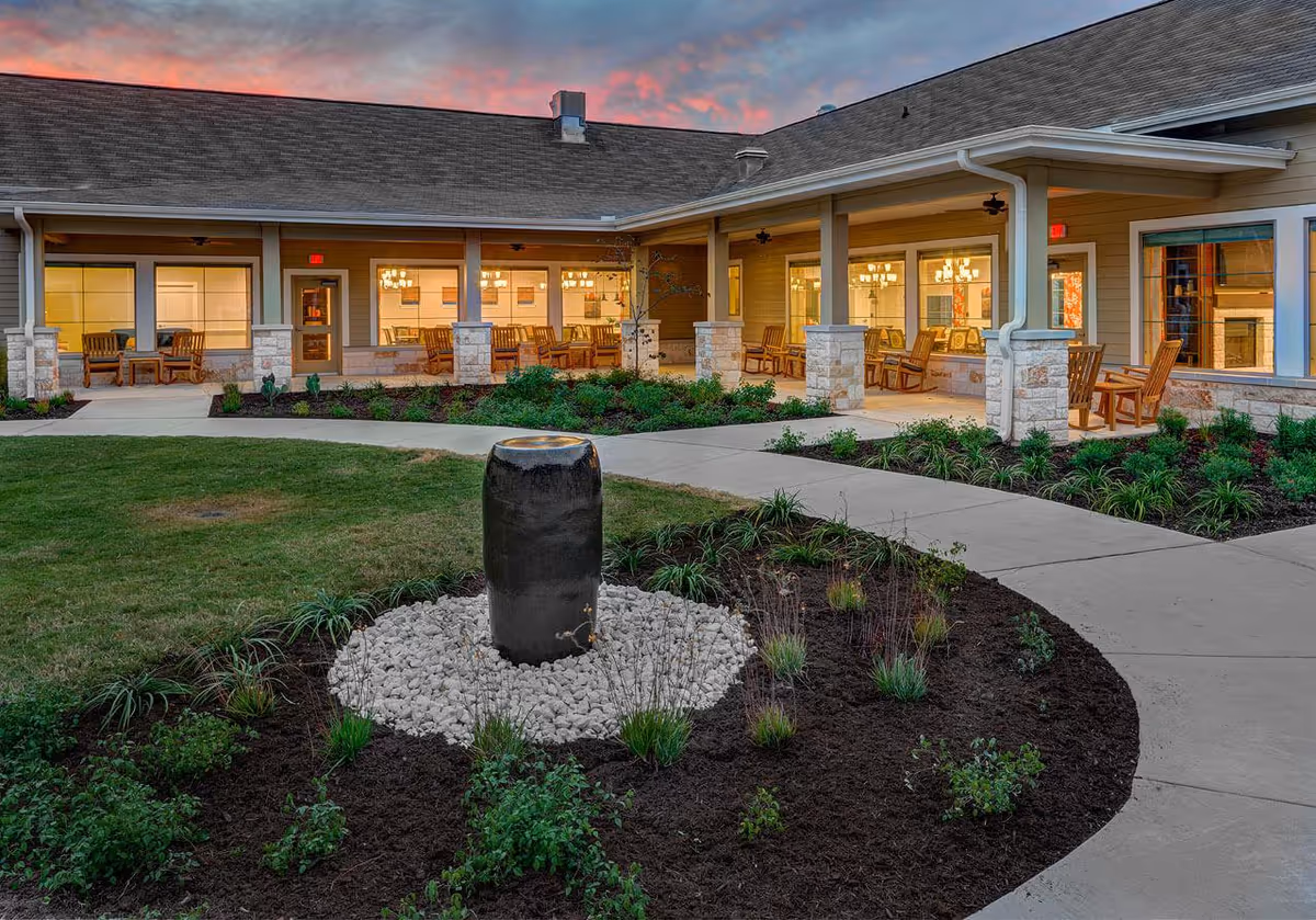 Outdoor courtyard area of The Delaney at Georgetown Village with a central water feature surrounded by landscaping and a paved walkway. The building has a covered porch with wooden rocking chairs and large windows showing a warmly lit interior at sunset.