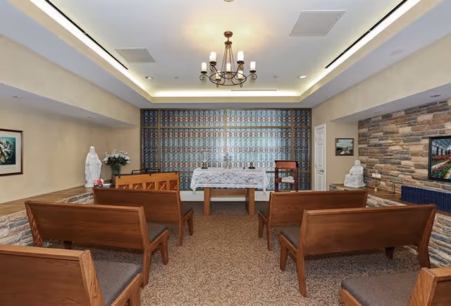 Interior of a small chapel or prayer room with wooden pews arranged facing an altar covered with a white cloth. The room has a chandelier hanging from the ceiling, stone accent walls, religious statues, and framed artwork on the walls.