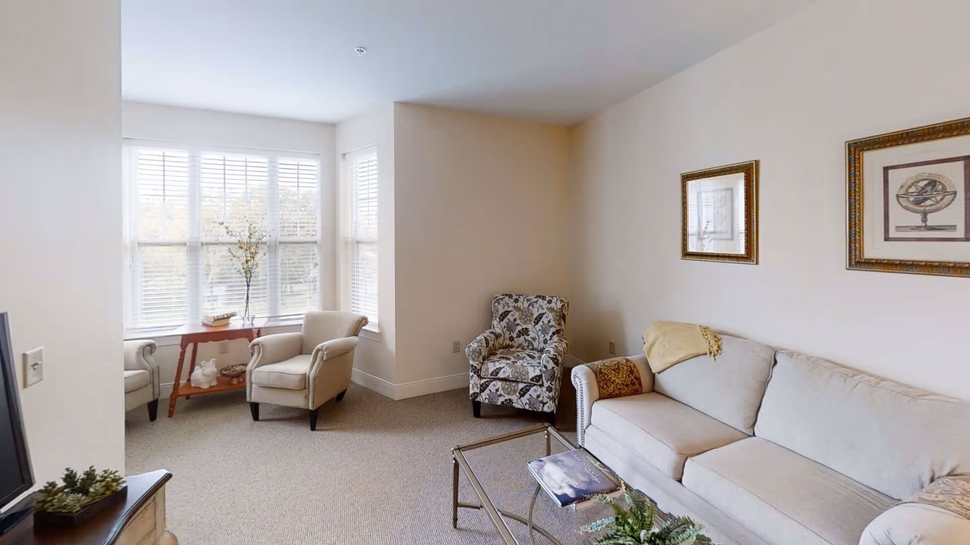 A bright living room with a beige sofa, a patterned armchair, two beige armchairs near a window with white blinds, a glass coffee table with a book and a plant, and framed artwork on the wall.