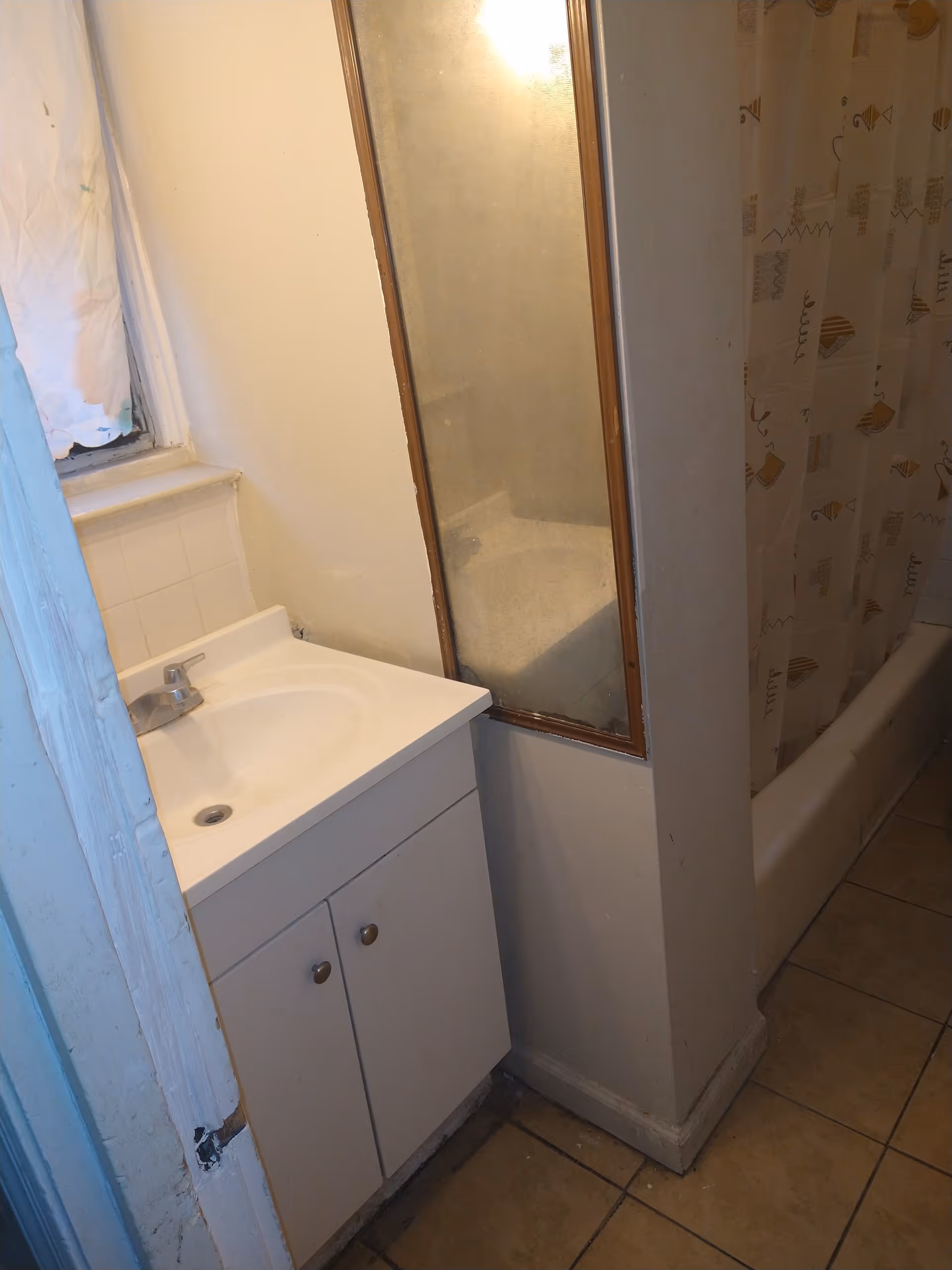 Small bathroom with a white sink cabinet, a faucet, a mirror with a wooden frame, a bathtub with a patterned shower curtain, and tiled floor. The bathroom appears to be compact and has a window with a curtain on the left side.