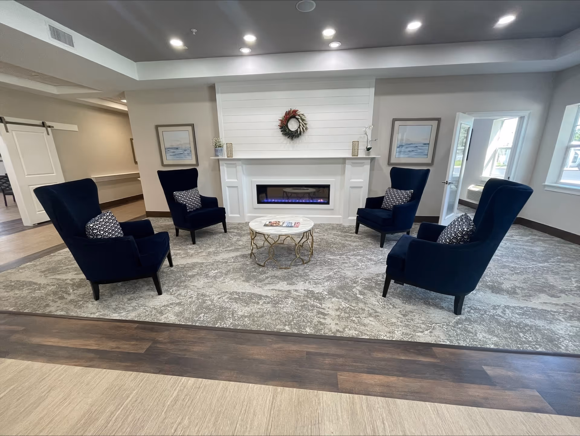 A cozy seating area in a senior living facility featuring four dark blue armchairs arranged around a round coffee table with magazines on it. The chairs are positioned on a large patterned rug in front of a white fireplace with a decorative wreath above it. The room has light-colored walls, recessed ceiling lights, framed artwork on either side of the fireplace, and windows letting in natural light.