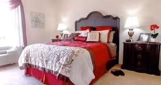 A cozy bedroom with a large bed featuring a dark wooden headboard, red and white bedding, and multiple pillows. There are two bedside tables with lamps, a framed photo, and a red flower on the right table. A window with sheer curtains is on the left side, and a pair of slippers is on the carpeted floor near the right bedside table.