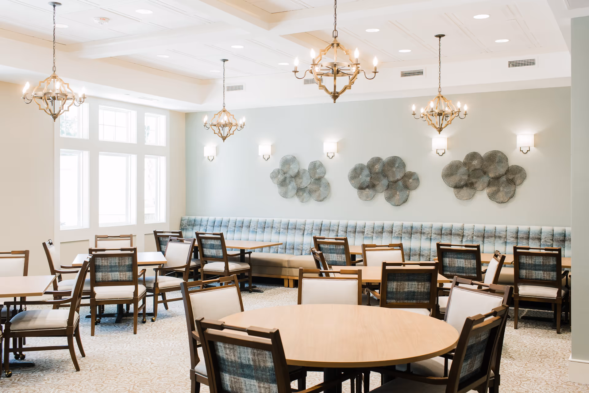 A bright dining room with multiple wooden tables and chairs arranged neatly. The room features large windows allowing natural light to fill the space, decorative wall art, and elegant chandeliers hanging from the ceiling. There is a long upholstered bench along one wall.