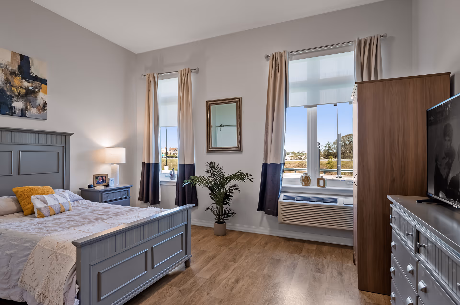 Bright furnished bedroom with a gray bed, nightstand, dresser and TV, two windows with curtains, and a potted plant.