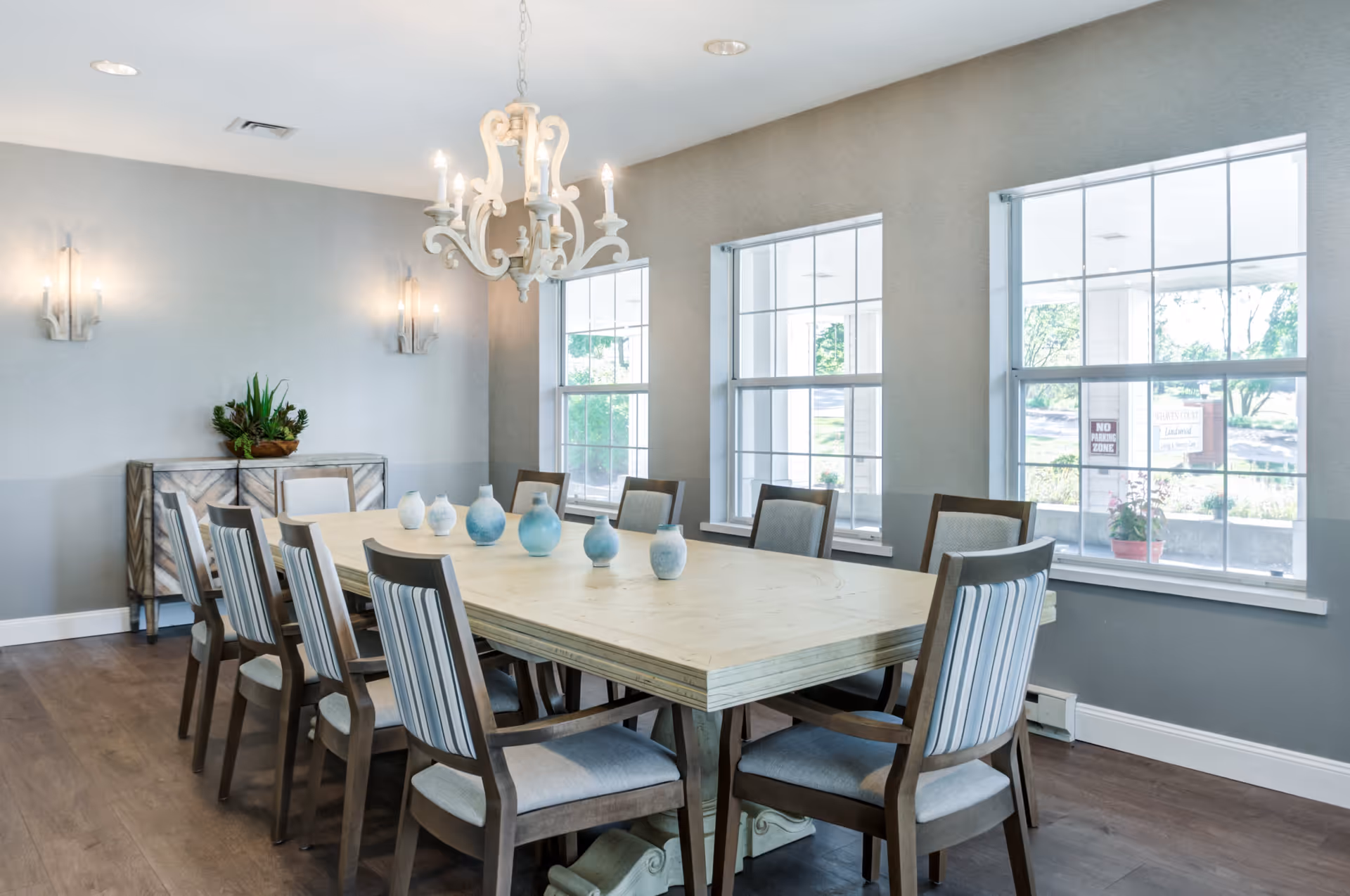A bright dining room with a long rectangular wooden table surrounded by eight cushioned chairs with striped backs. The room has three large windows letting in natural light, a white chandelier hanging above the table, and a sideboard with a plant on it against the wall.