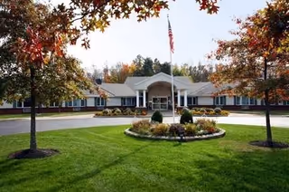 Front exterior view of Vinecroft Retirement Community building with a circular driveway, a flagpole with an American flag, well-maintained green lawn, and trees with autumn-colored leaves.