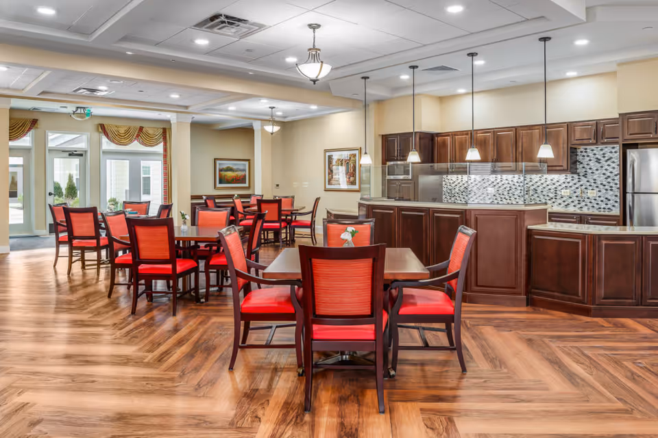 Communal dining room with red-upholstered chairs around wooden tables and a dark wood kitchen and service counter in the background.