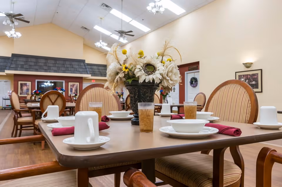 A dining room in an assisted living facility with tables set for a meal. The tables have white cups, bowls, plates, glasses of iced tea, and red cloth napkins. There is a large floral centerpiece on the table in the foreground. The room has high ceilings with ceiling fans and chandeliers, and several framed pictures on the walls.