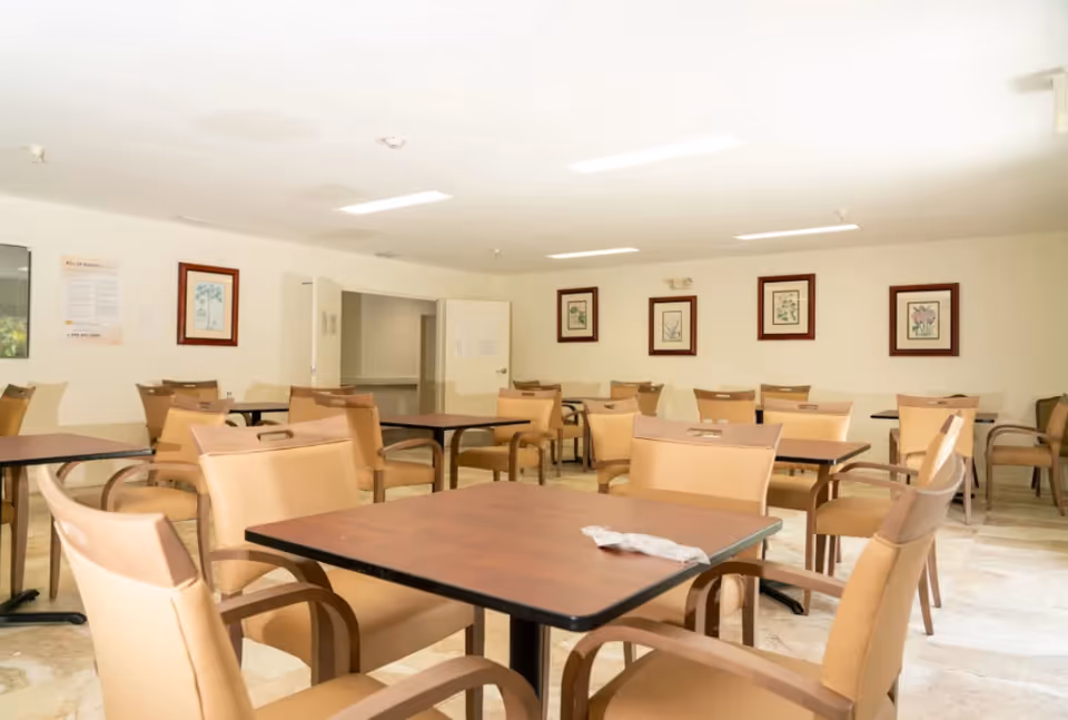 A bright dining room with multiple square wooden tables and tan cushioned chairs arranged neatly. The walls are white and decorated with framed botanical prints. There is an open doorway leading to another room.