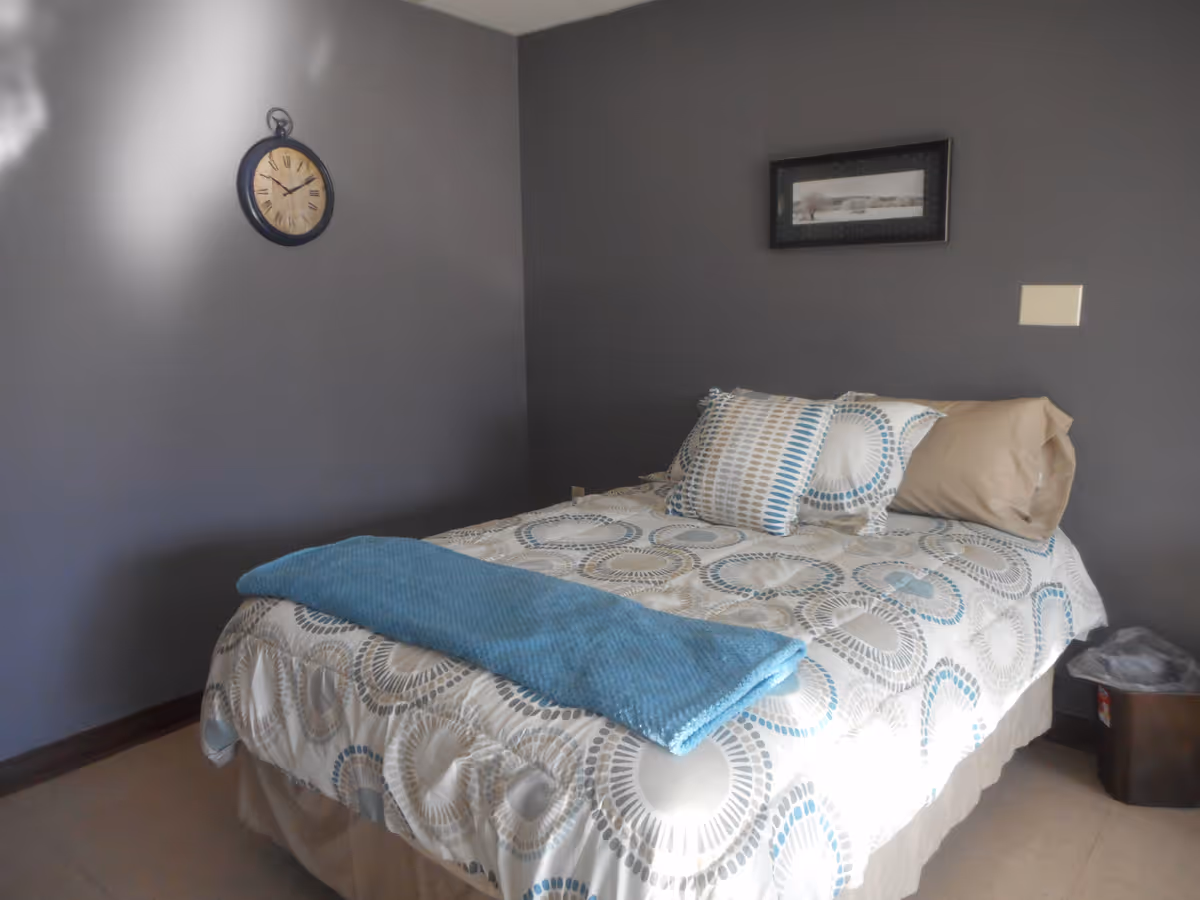 Small bedroom with a single bed against a gray wall, patterned bedding and a blue throw, with a wall clock and framed picture above.