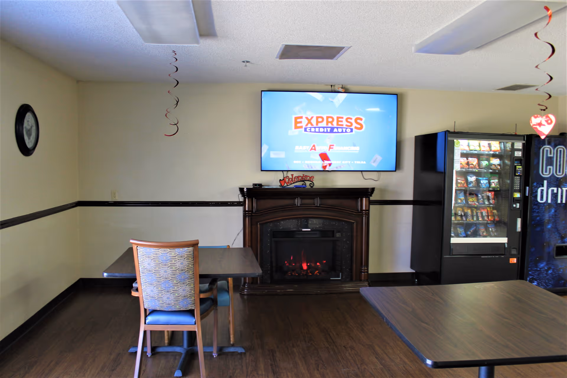 A small common area with two tables and chairs, a wall-mounted TV displaying an advertisement for Express Credit Auto, an electric fireplace below the TV, a vending machine with snacks, and a soda vending machine. The room has light-colored walls with a dark chair rail and dark wood flooring. Red spiral decorations hang from the ceiling.