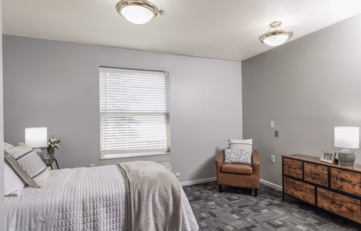 A cozy bedroom with a neatly made bed covered in white bedding and a light gray blanket. There is a small bedside table with a lamp and a vase of flowers next to the bed. A window with white blinds is centered on the wall behind the bed. Across from the bed, there is a brown leather armchair with a decorative pillow and a wooden dresser with a lamp and framed photos on top. The walls are painted light gray and the floor is carpeted with a patterned design.