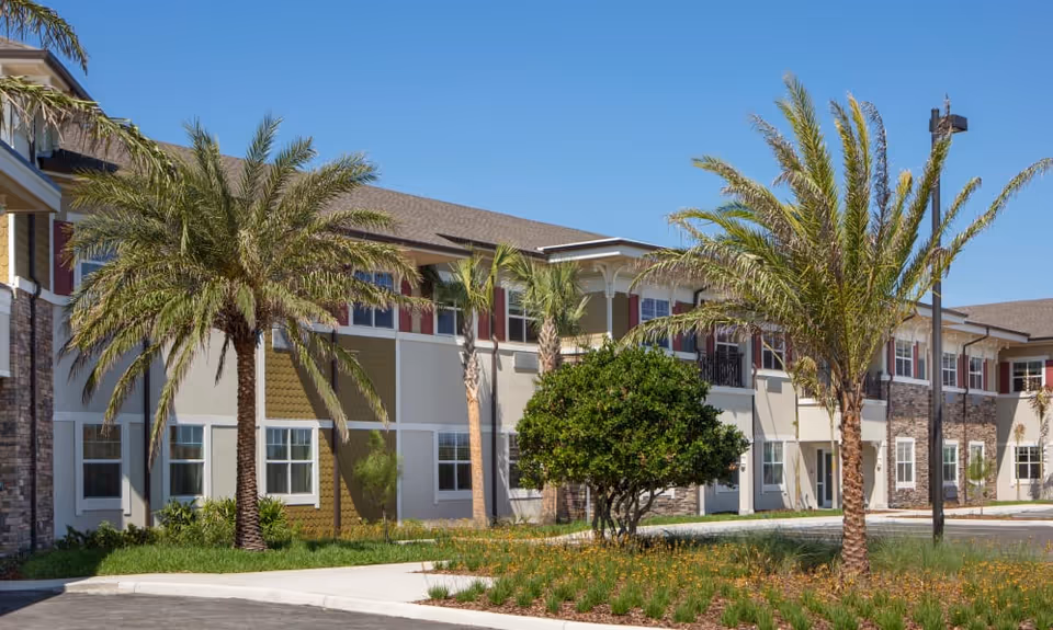 Exterior view of a two-story assisted living facility building with palm trees and landscaping in front under a clear blue sky.