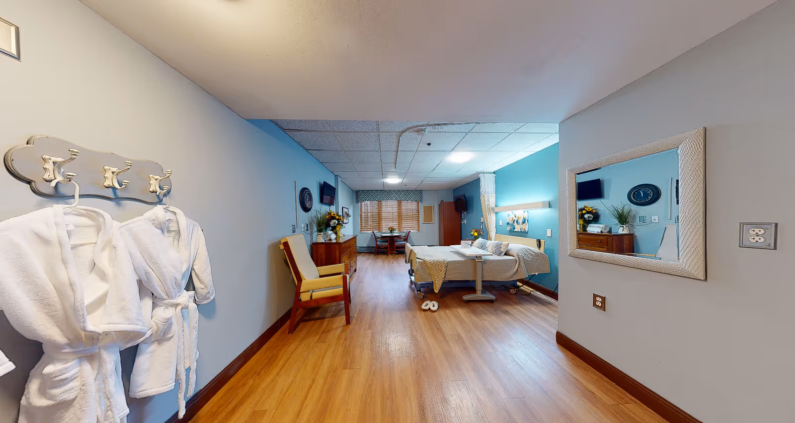 A spacious and well-lit rehabilitation room featuring a hospital bed with side rails, a bedside table, and a comfortable chair. The room has wooden flooring, light blue and gray walls, a large mirror on one wall, and two white bathrobes hanging on hooks. In the background, there is a small dining table with chairs near a window with blinds, and a dresser with flowers and a TV mounted on the wall.