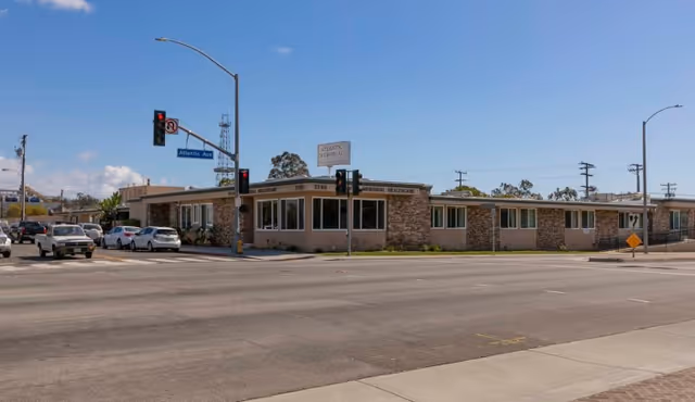 Exterior view of Atlantic Memorial Healthcare Center, a single-story building with large windows and stone accents, located at a street corner with traffic lights and several cars waiting at the intersection under a clear blue sky.