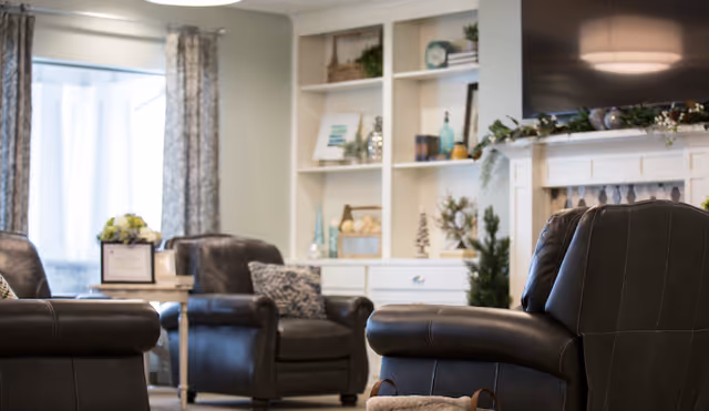 A cozy living room area with dark leather armchairs arranged around a small table. In the background, there is a white built-in shelving unit decorated with various ornaments and plants, and a large flat-screen TV mounted above a white fireplace mantel. Light filters in through a window with patterned curtains.
