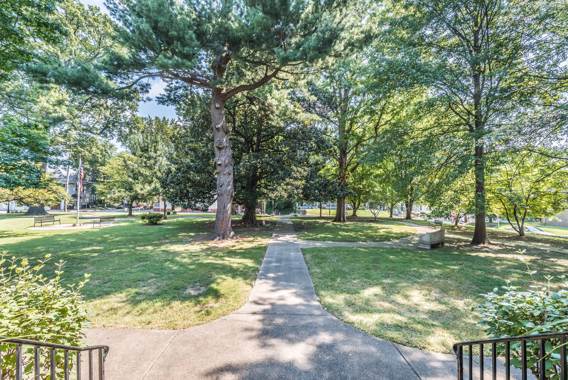 A sunny outdoor park area with a concrete pathway leading through green grass and several large trees providing shade. Benches are placed along the path and an American flag is visible in the background.