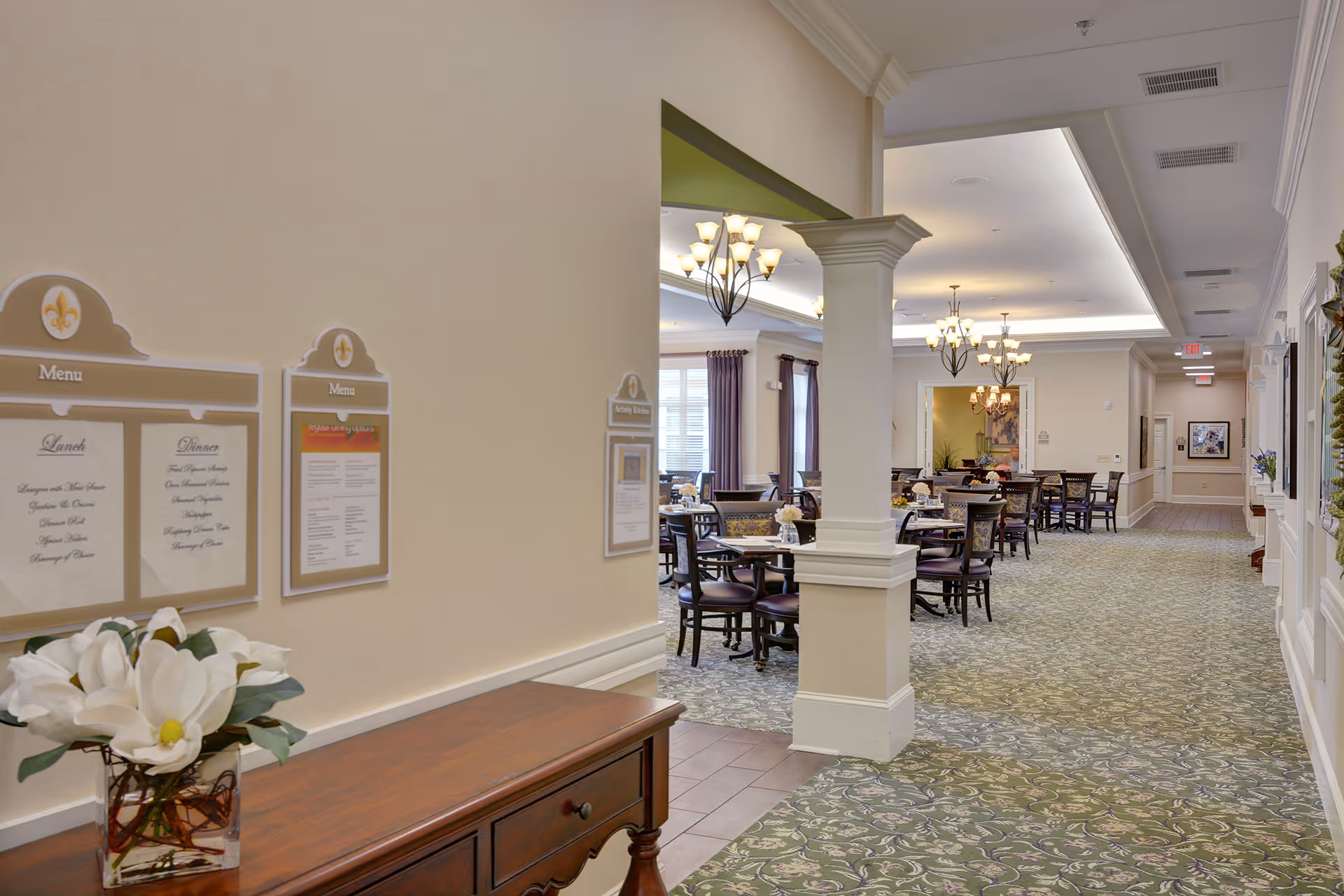 A spacious dining room with multiple round tables and chairs, chandeliers, and menus displayed on a wall.