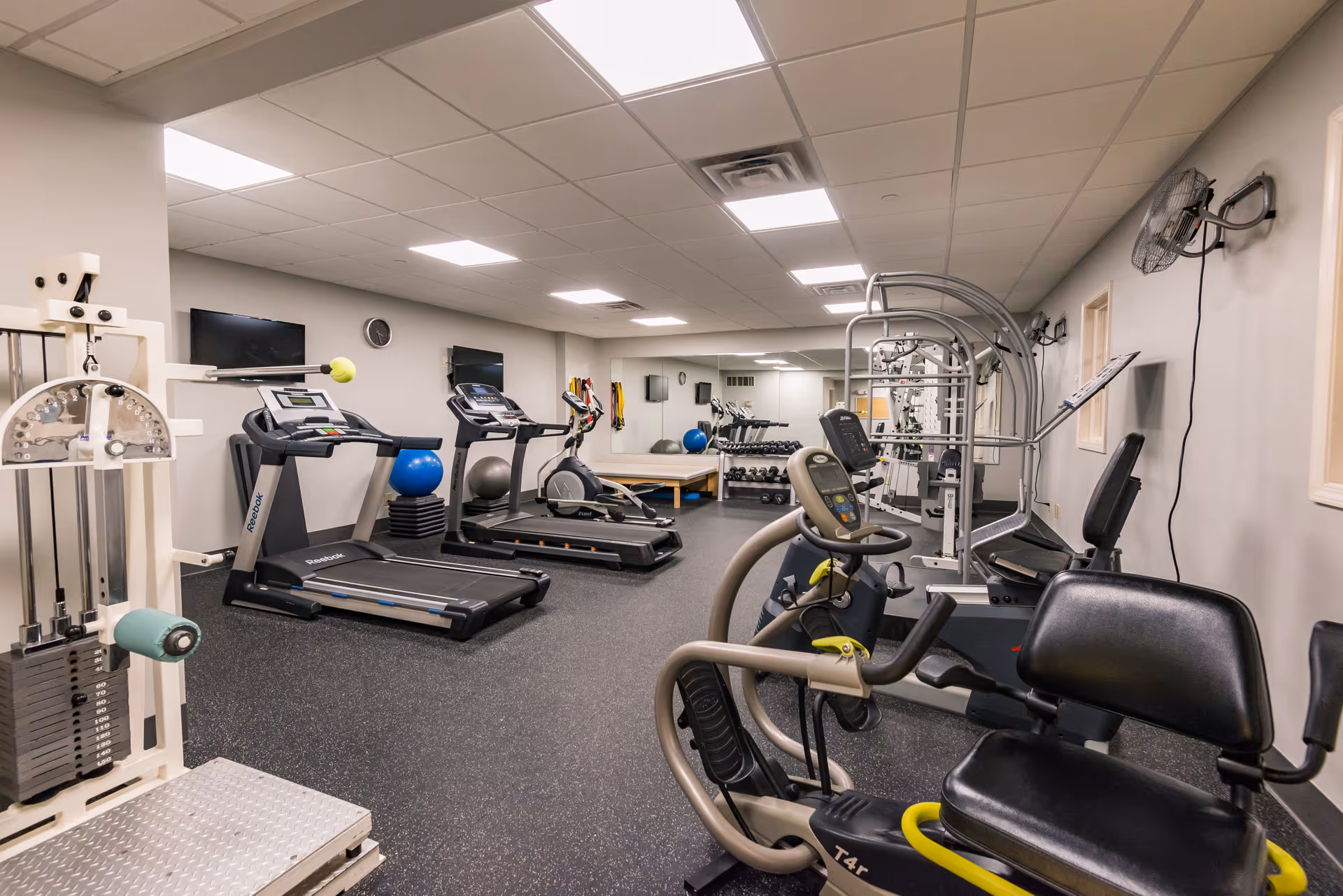 Interior view of a fitness room with various exercise equipment including treadmills, stationary bikes, weight machines, and free weights. The room has a gray floor, light gray walls, ceiling lights, wall-mounted fans, and mirrors along one wall.