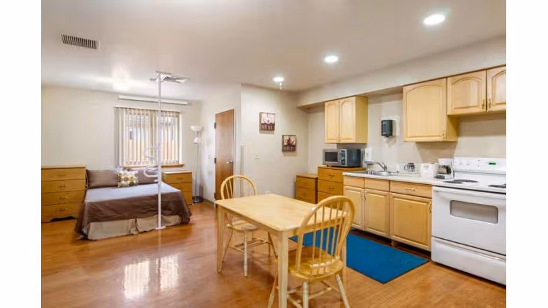 A combined living space featuring a bedroom area with a bed, dresser, and a ceiling lift, adjacent to a kitchen area with wooden cabinets, a microwave, a stove, and a small dining table with two chairs. The room has wooden flooring and neutral-colored walls.