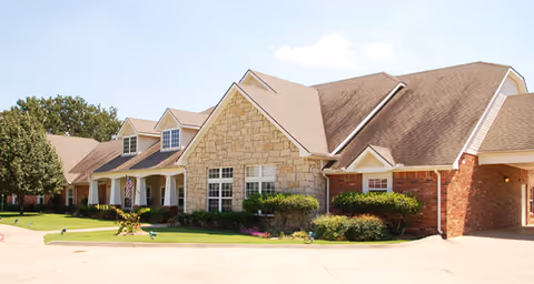 Exterior view of a single-story senior living facility building with a combination of stone and brick facade, multiple gabled roofs, well-maintained landscaping, and a clear blue sky.