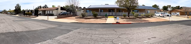 Panoramic view of a single-story residential building with a beige exterior, surrounded by a dry landscape with sparse trees and shrubs. Several cars are parked along the driveway and street in front of the building under a clear sky.