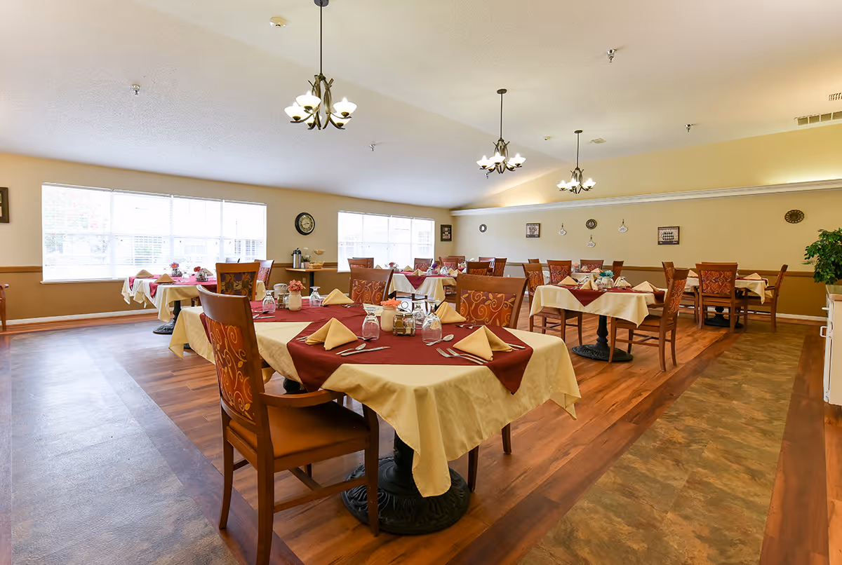 A bright dining room with multiple tables covered in cream tablecloths and burgundy runners, each set with folded napkins, glasses, and silverware. The room has wooden chairs with patterned upholstery, large windows letting in natural light, and decorative wall clocks and pictures. Three chandeliers hang from the ceiling, and the floor has a combination of wood and tile patterns.