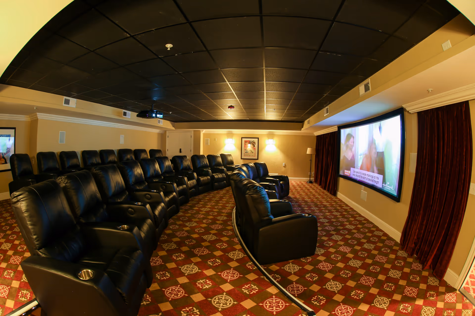 A cozy theater room with two rows of black leather recliner chairs facing a large screen on the wall. The room has a patterned carpet with red and beige tones, beige walls, and a black tiled ceiling. There are wall sconces providing warm lighting and a projector mounted on the ceiling.