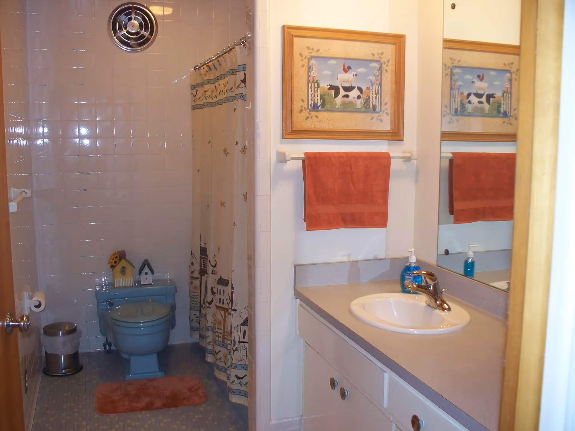 A bathroom with a blue toilet, a shower with a curtain decorated with houses and flowers, a white sink with a silver faucet on a beige countertop, a red towel hanging on a towel rack, a framed picture of farm animals above the towel, and a small trash can next to the toilet.