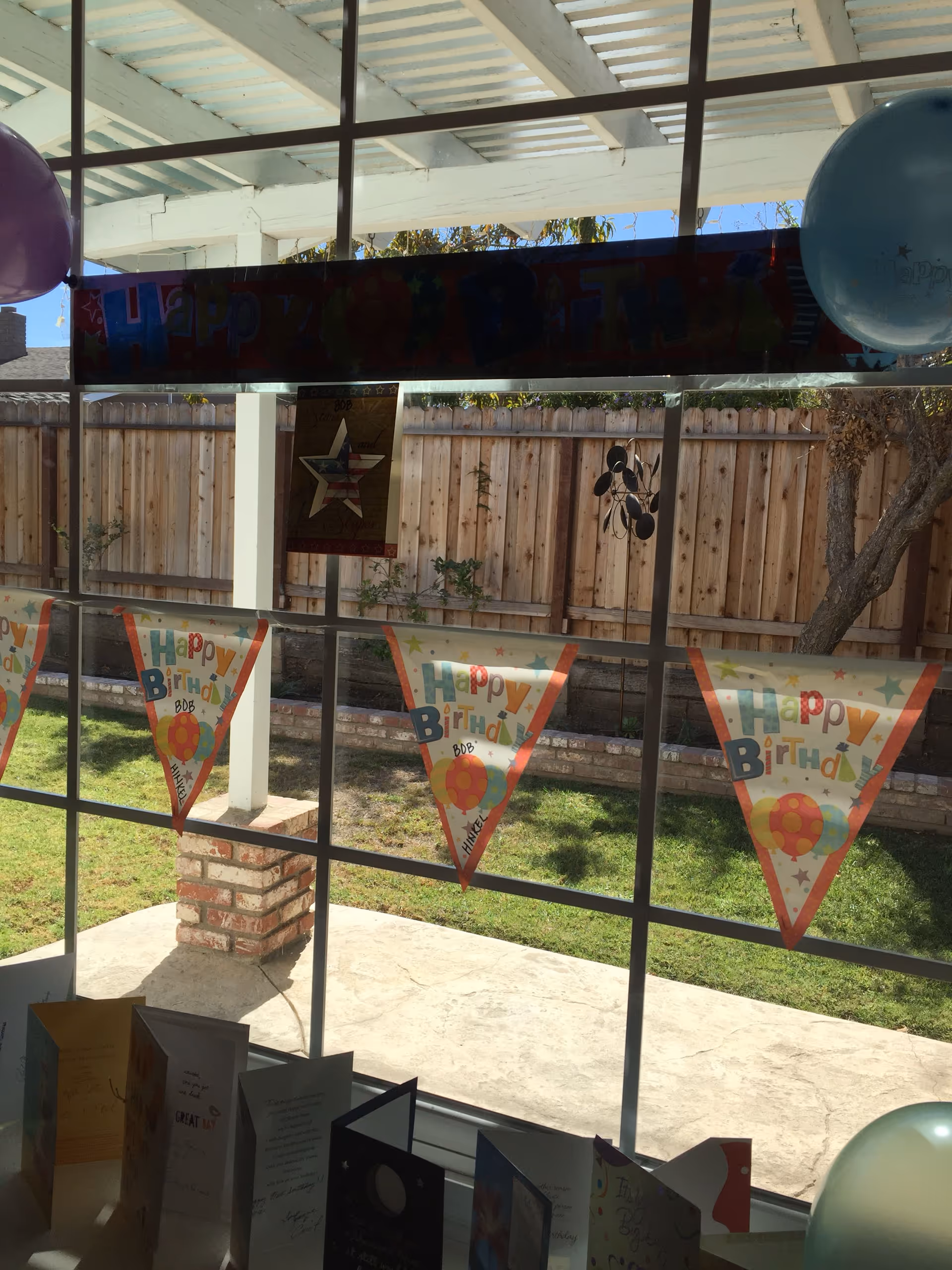 View through a window showing a backyard with a wooden fence and a covered patio area. Birthday decorations including a 'Happy Birthday' banner, triangular flags with 'Happy Birthday Bob' written on them, and balloons are visible inside near the window. Several birthday cards are displayed on a surface below the window.