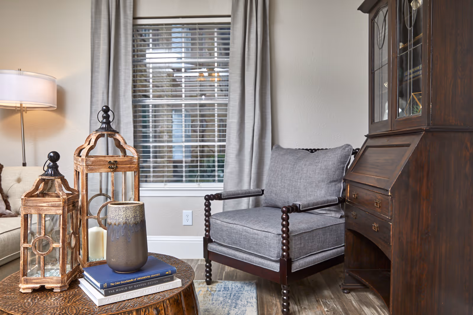 A cozy living room corner featuring a wooden side table with decorative lanterns and a ceramic vase on top of stacked books, a gray cushioned armchair with wooden frame, a wooden cabinet with glass doors, a window with blinds and gray curtains, and a floor lamp in the background.