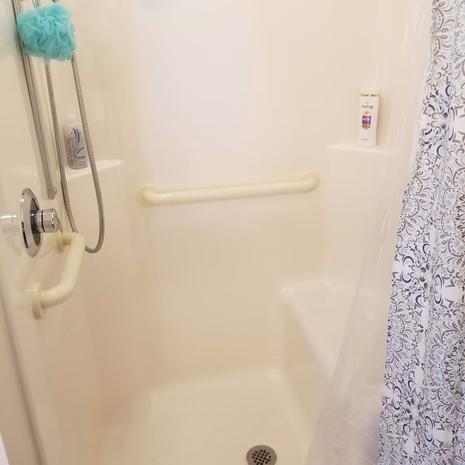 A white shower stall with a built-in seat and two grab bars for support. There is a handheld showerhead with a silver hose hanging on the left side, a blue bath pouf hanging from the showerhead, a bottle of lavender-scented body wash on a small shelf, and a bottle of Pantene shampoo on another shelf. A patterned shower curtain with blue and gray designs is partially visible on the right side.