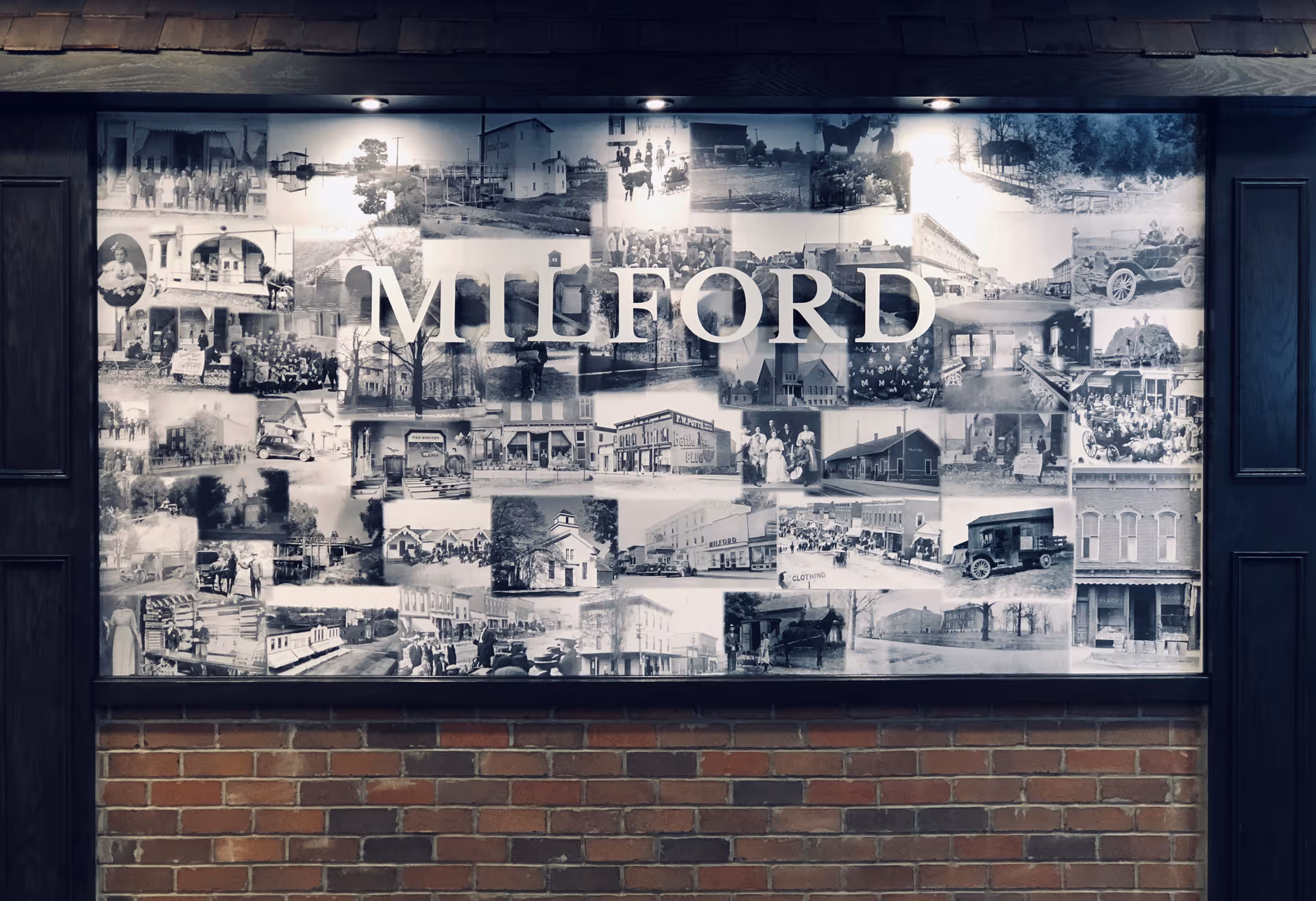 A large collage of black and white historical photographs displayed on a wall with the word 'MILFORD' prominently overlaid in large white letters. The collage is framed by dark wood panels and illuminated by overhead lights, with a brick wall below.
