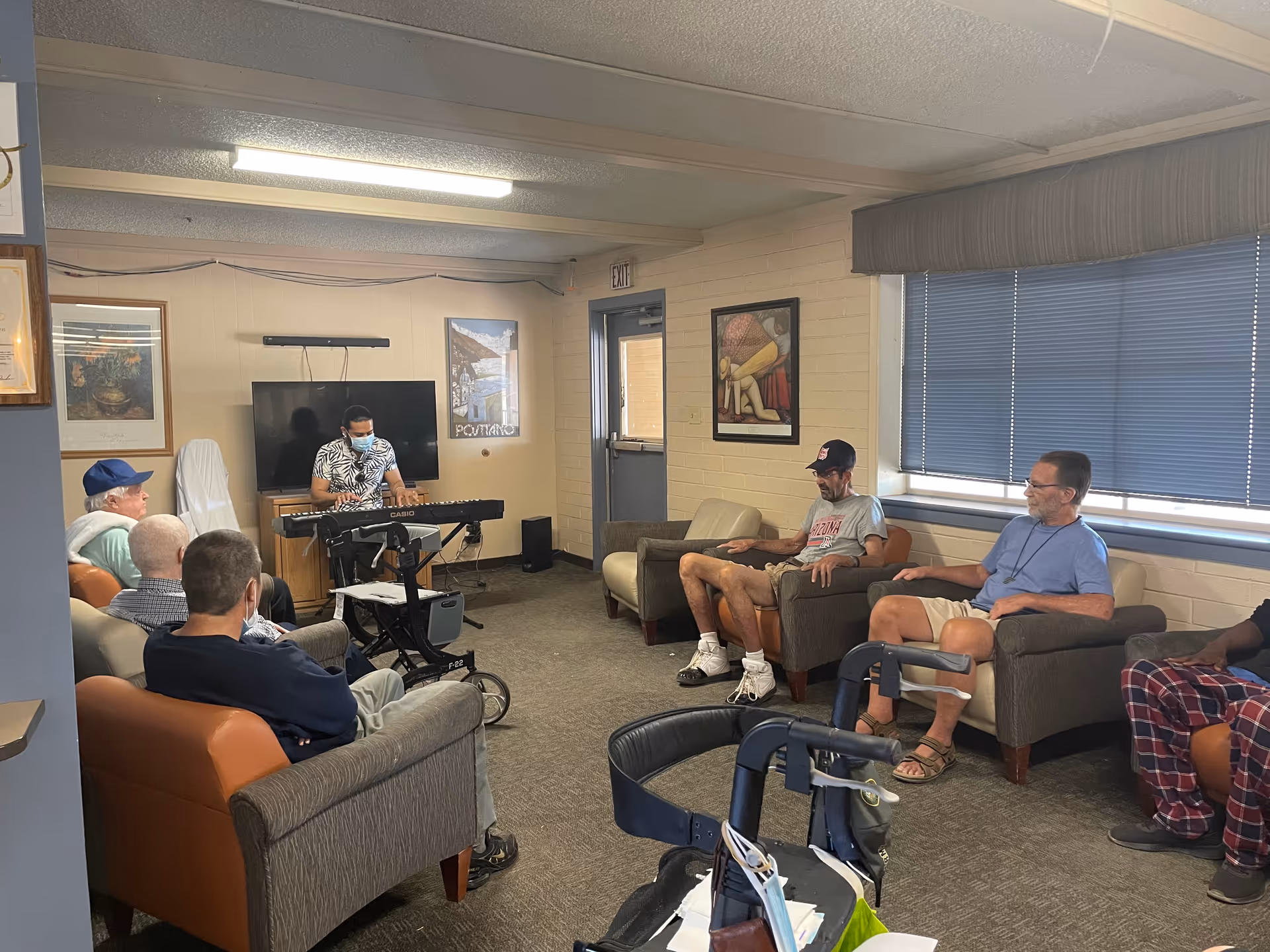 A common lounge room with several men seated in chairs while a masked musician plays an electronic keyboard.