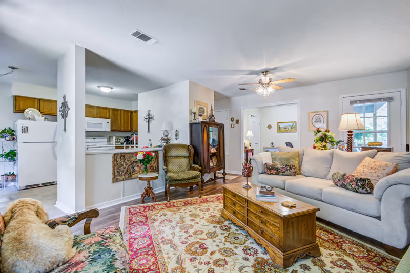 A cozy living room in a retirement community featuring a beige sofa with floral cushions, a wooden coffee table with books and decorative items, an armchair with a floral pattern, and a green upholstered chair. The room has a ceiling fan with lights, a patterned area rug, and a glass-front cabinet. The kitchen with wooden cabinets and white appliances is visible in the background. A dog is lying on the floral armchair.